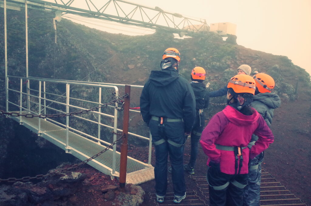 Photo of people on a misty day with helmets and jackets about to step onto a platform to descend into a Volcano in Iceland