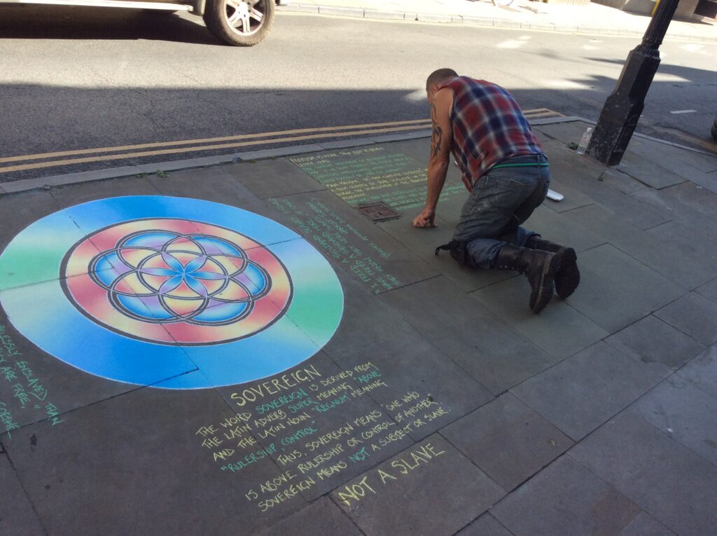 My photograph ofA man deals on the floor with chalk drawing and writing images on the floor in Glastonbury. You can see a sacred geometry drawing and below that that’s writing.