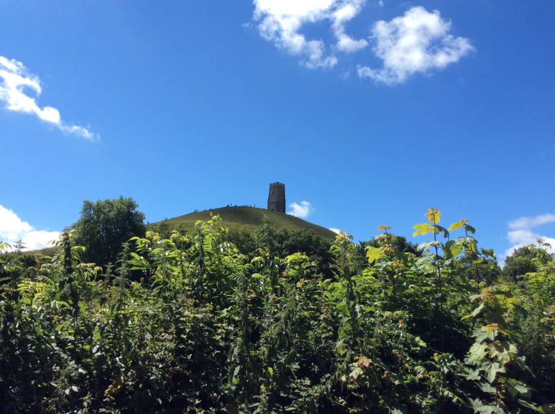 My photograph of Glastonbury Tor from a distance (below the Tor) with a vivid blue sky with faint clouds and vibrant foliage below.