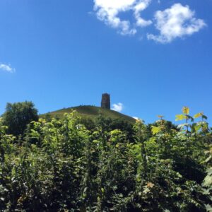 My photograph of Glastonbury Tor from a distance (below the Tor) with a vivid blue sky with faint clouds and vibrant foliage below.