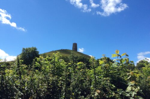 My photograph of Glastonbury Tor from a distance (below the Tor) with a vivid blue sky with faint clouds and vibrant foliage below.