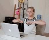 A moving image of a woman at her desk with earphones watching her MacBook and doing exercise.