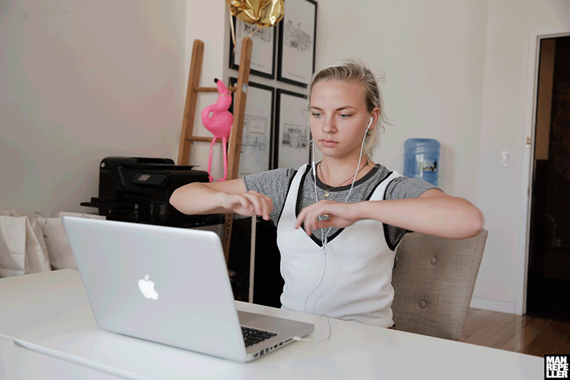 A moving image of a woman at her desk with earphones watching her MacBook and doing exercise.