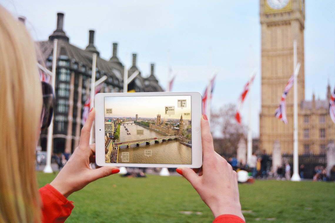 A photoshopped potography iPad Mini in the hands of a woman outdoors the house of Parliment the image in the ipad is of another side of Parliment near the river with captions of where I was born