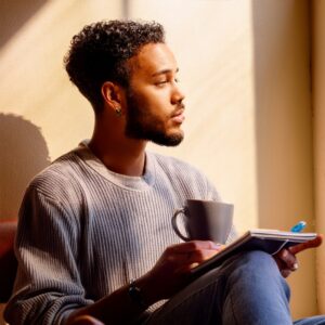 A man seated, hands holding a small object ( a cup and notebook), head his eyes focused on something just out of frame. Environment: Calm, minimalistic space; soft shadows; natural muted light. a shadow or reflection of himself in the background to symbolize self-awareness and a Aqaurius man with his moon in Virgo