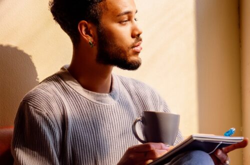 A man seated, hands holding a small object ( a cup and notebook), head his eyes focused on something just out of frame. Environment: Calm, minimalistic space; soft shadows; natural muted light. a shadow or reflection of himself in the background to symbolize self-awareness and a Aqaurius man with his moon in Virgo